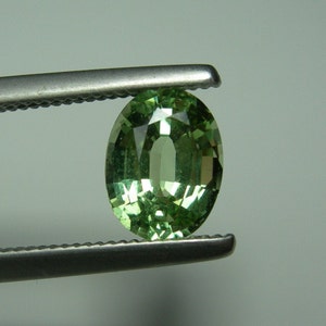 Può includere: Una pietra preziosa di colore verde chiaro con taglio ovale e superficie sfaccettata, tenuta da una pinzetta metallica color argento. La pietra mostra riflessi interni e una qualità traslucida, esibendo un colore verde lime brillante.