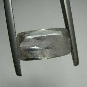 May include: A clear, faceted gemstone held by metal tweezers. The gem has a rectangular shape with cut edges and a slightly curved top. The stone appears translucent with internal inclusions, reflecting light. The background is a neutral gray.
