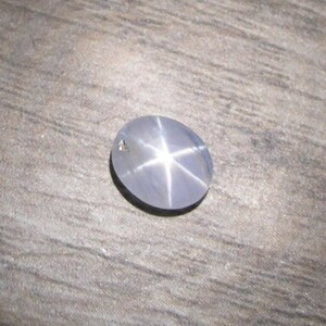 May include: A polished, oval-shaped grey star sapphire gemstone featuring a distinct six-rayed star pattern centered on its surface. The stone exhibits a smooth, cabochon cut with a translucent appearance and a small, dark inclusion near the edge.