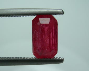 Puede incluir: Un rubí rectangular facetado, sostenido por unas pinzas plateadas. El rubí es de un color rojo intenso y vibrante, con estructuras internas visibles. Las pinzas están enfocadas, resaltando la claridad y el corte de la gema.