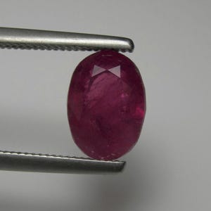 May include: A faceted, oval-shaped ruby gemstone held by tweezers. The ruby is a deep, rich red colour with a polished surface. The gemstone is a focal point, showcasing its clarity and brilliance.