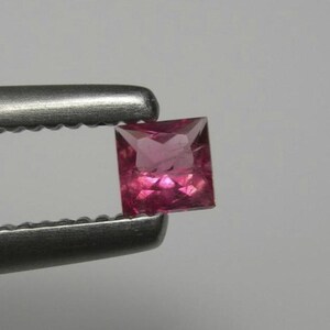 May include: A square-cut, faceted gemstone with a vibrant magenta-pink hue. The stone is held by a pair of metallic, serrated-edge tweezers, showcasing its transparent quality and precise geometric shape against a neutral background.