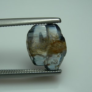 May include: A rough-cut, blue-grey gemstone held by a pair of tweezers. The gem has a natural, unpolished appearance with areas of brown and tan. The stone's surface reflects light, showcasing its crystalline structure. The tweezers are silver.