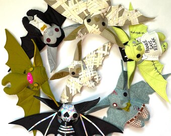 Könnte beinhalten: Eine Sammlung handgefertigter Plüschfledermäuse in verschiedenen Farben und Designs. Einige Fledermäuse sind schwarz, grün und grau, mit Details wie Mumienwicklungen, Skelettmustern und dem Text "Frankie Says Relax". Diese Halloween-Dekorationen sind kreisförmig angeordnet.