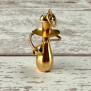 May include: A gold-colored charm in the shape of a hookah. The charm has a round base, a cylindrical body, and a small, round top. A coiled rope-like design wraps around the body. A small ring is attached to the top for hanging.