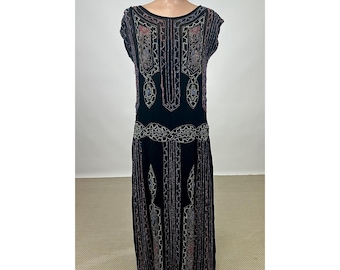 May include: A black sleeveless dress featuring intricate beadwork in silver, red, and blue tones. The garment displays a geometric pattern across the bodice and skirt, with a dropped waistline and a straight silhouette that extends to the floor.