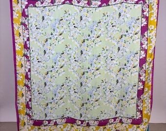 Op de afbeelding: Een vierkante sjaal met een bloemenpatroon. Het midden is lichtgroen met witte en blauwe bloemen. Het heeft een paarse rand, een zwarte rand en een gele buitenrand met witte bloemen. De sjaal wordt gepresenteerd op een blauw rolrek.