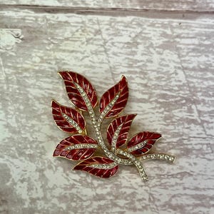Op de afbeelding: Een rode emaille en strass blad broche. De bladeren zijn in een cluster gerangschikt en hebben een gouden afwerking.