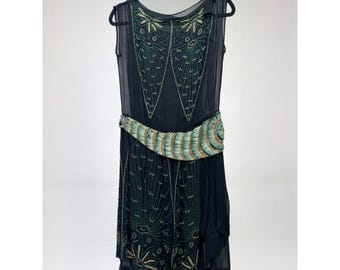 May include: Black sleeveless dress with intricate green and gold beaded embellishments. The dress features a decorative belt with teal and gold accents. The dress is hanging on a black hanger.