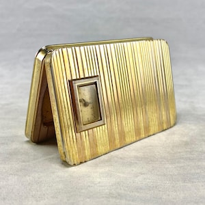 May include: A vintage gold-toned travel clock with a rectangular shape and vertical ribbed detailing. The clock face is small and square, set within a gold frame. The clock is open, revealing the clock face.