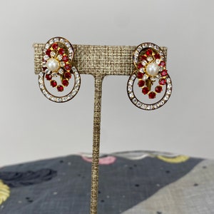 May include: A pair of gold tone clip-on earrings with a red and clear rhinestone design. Each earring features a single pearl in the center.