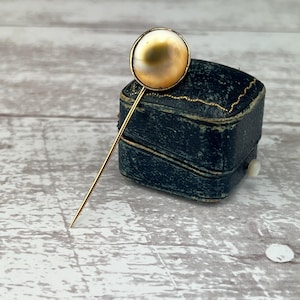 Puede incluir: Un broche de corbata de oro con una piedra redonda e iridiscente similar a una perla.