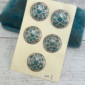 May include: Five round, silver-colored buttons with a turquoise floral design. The buttons are arranged in a grid pattern on a white card with the text "No. 3" written in black ink.