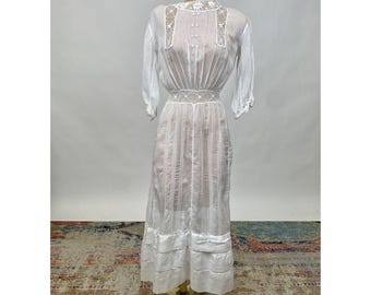 May include: A white vintage dress with lace detailing and a gathered skirt. The dress has a high neckline and three-quarter sleeves.