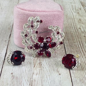 May include: A silver brooch with a floral design featuring red and clear rhinestones. The brooch is displayed on a pink velvet cushion. Two matching earrings with red rhinestones are also included.