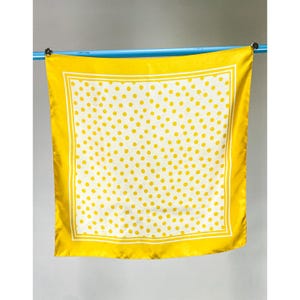 May include: A square scarf featuring a white center with a yellow polka dot pattern, framed by a solid yellow border and a thin white line. The fabric has a smooth, satin-like finish and is displayed hanging from a blue horizontal rod.