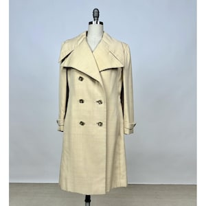 May include: A double-breasted beige coat featuring a wide notched collar, long sleeves with cuff straps, and six dark buttons arranged in two vertical rows. The garment is displayed on a mannequin, showcasing a structured silhouette and textured fabric.