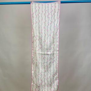 May include: A white silk scarf with a pink floral pattern. The scarf has a pink border and is hanging on a blue rod.