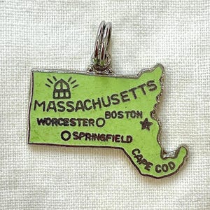 May include: A silver-toned charm in the shape of the state of Massachusetts. The charm is green with the state name and city names in black. The charm includes a silver loop for attaching to a bracelet or necklace.