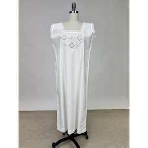 Puede incluir: Un camisón blanco, largo hasta el suelo, sobre un maniquí. El escote cuadrado y las mangas cortas están ribeteados con encaje. El corpiño presenta diseños florales bordados. El camisón está hecho de una tela lisa, posiblemente satinada.