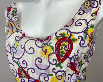 Op de afbeelding: Een witte, mouwloze jurk met een kleurrijk bloemenpatroon. Het patroon bevat swirls, paisley-vormen en rode, gele, groene en paarse bloemen.
