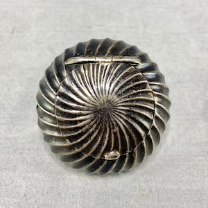 Puede incluir: Un estuche compacto redondo de metal oscuro con un patrón prominente en espiral similar a un rayo de sol que irradia desde el centro. La superficie tiene un diseño acanalado y texturizado con una tapa con bisagras y un pequeño cierre.
