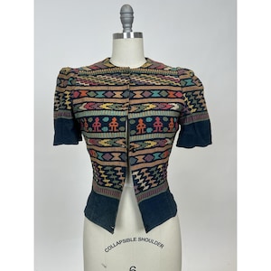 May include: A vibrant, open-front jacket with short sleeves. The jacket showcases a detailed, geometric pattern in shades of blue, orange, yellow, and green. The sleeves and lower section of the jacket are a solid dark blue. The jacket is displayed on a mannequin.