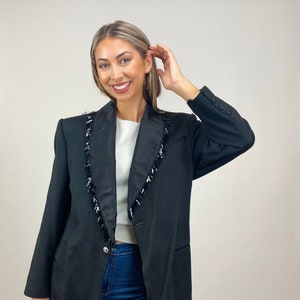 May include: A black blazer with a satin collar and a fringe trim. The blazer has a single button closure and two pockets.