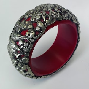 Pode incluir: Um bracelete vermelho em forma de bangle com um design de filigrana prateada que apresenta intrincados motivos florais e de pássaros.