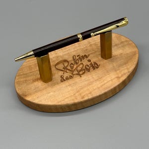 May include: A brown and gold pen with gold accents is displayed on a wooden pen holder. The holder is oval shaped and has the words "Robin Sas Bau" engraved on it.