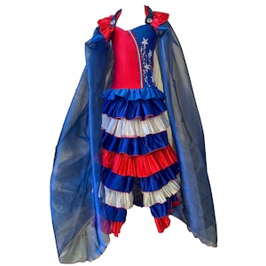 Puede incluir: Un vestido rojo, blanco y azul con falda de volantes y una capa a juego. El vestido tiene un corpiño con un diseño de estrella y fuegos artificiales.