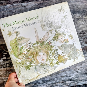 Peut inclure: Un livre ancien intitulé "The Magic Island" de Janet Marsh. La couverture présente une illustration fantaisiste avec un visage d'enfant entouré de feuillage, un lézard, un hérisson et d'autres éléments fantastiques. Le titre est en vert.