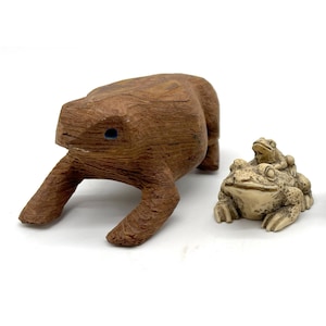 May include: Four animal figurines: a carved wooden frog, a beige frog with a smaller frog on its back, a black stone Foo Dog, and a brown turtle. The figurines are on a white surface.