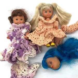 May include: Three crocheted doll outfits in pink, purple, and white. The outfits are made with a delicate, lacy stitch. The dolls are not included.