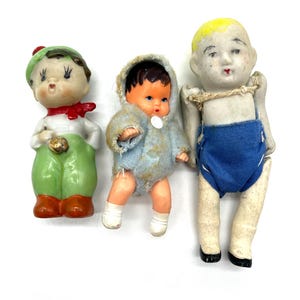 Könnte beinhalten: Drei Porzellanpuppen, jede mit einem anderen Outfit. Die Puppe links trägt ein grünes und rotes Outfit, die Puppe in der Mitte trägt ein blaues Kapuzenoutfit und die Puppe rechts trägt eine blaue Shorts.