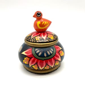 May include: A small, round wooden box with a bird finial. The box is painted with a floral design in shades of red, orange, yellow, green, and blue on a black background. The lid of the box has a gold trim.