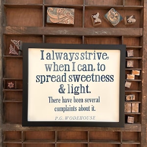May include: A framed print with the quote "I always strive, when I can, to spread sweetness & light." in dark blue text. The print is set against a wooden background with small, decorative blocks.