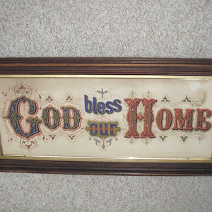 May include: A framed print with the words "God bless our Home" in ornate, colorful lettering. The frame is dark brown with a gold inner border, and the background is a light beige color.