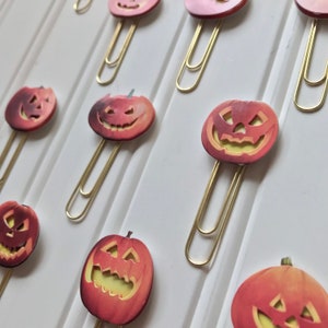 May include: A set of gold paper clips with orange and yellow jack-o'-lantern designs. The jack-o'-lanterns have a carved smile and eyes.