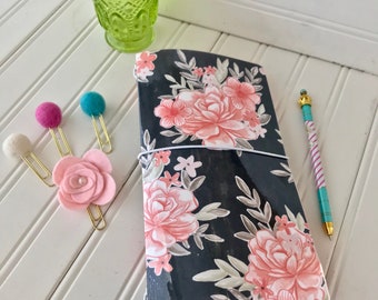 May include: A black and white floral patterned journal with a pink elastic band and a red edge. The journal is on a white surface with a green glass vase and flowers.