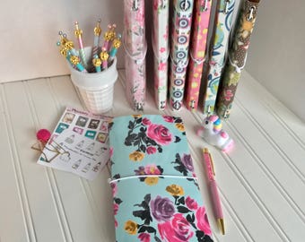 May include: A light blue floral patterned notebook with a white elastic band. The notebook is closed and sitting on a white surface. There are several colorful patterned notebooks in the background.