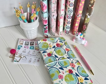 May include: A white and blue patterned journal with a globe design and a pink pen. The journal is closed and has a white elastic band around it. There are several colorful patterned journals in the background.