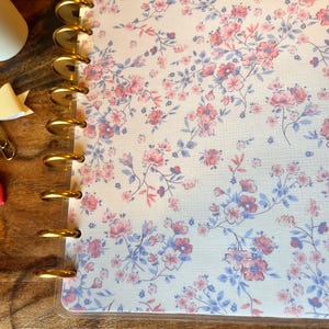 May include: A floral-patterned planner with gold rings and a clear cover. The design features pink and blue flowers on a white background. Decorative bow paper clips in pink and cream are also visible.