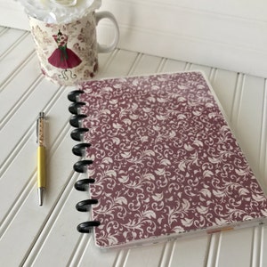 May include: A burgundy planner with a white floral pattern and a clear plastic cover. The planner has a black disc binding system and is lying on a white surface.