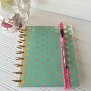 May include: A teal and gold polka dot planner with a pink ribbon bookmark and a black pen.