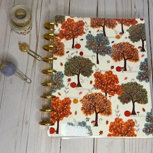 May include: A planner with a fall-themed cover featuring trees in shades of orange, green, and blue, along with pumpkins and leaves. The planner has gold-colored rings and is accompanied by decorative tape and paper clips.