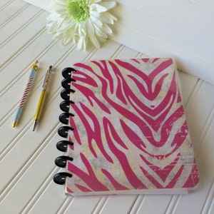 May include: A pink and white zebra print notebook with a black coil binding. The notebook is lying on a white surface with two pens next to it.