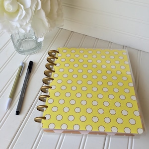 May include: A yellow polka dot planner with gold rings. The planner is closed and lying on a white surface.
