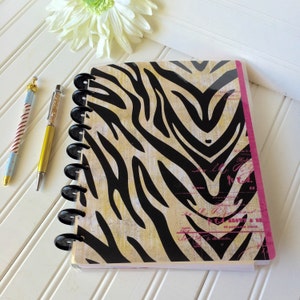 May include: A black and tan zebra print notebook with a pink edge and black coil binding. The notebook has a vintage style design with faded text and a handwritten style font.