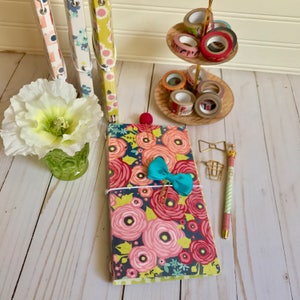 May include: A floral patterned journal with a turquoise bow, gold binder clips, and a gold pen with a striped design. The journal is on a white wooden surface with a decorative stand holding colorful washi tape.
